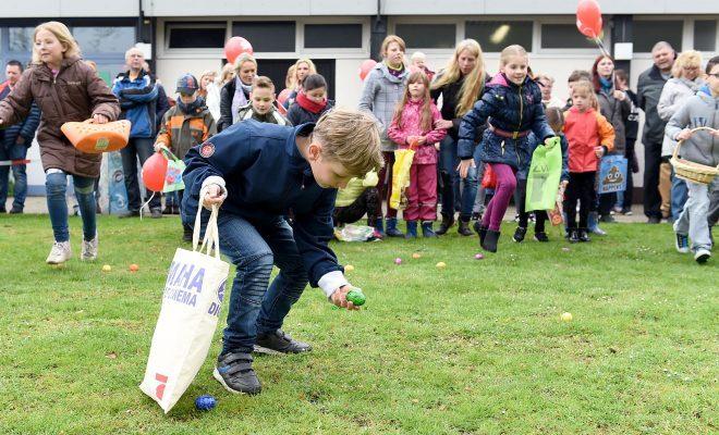 Ein Junge hebt ein buntes Ei vom Boden auf, während Kinder und Erwachsene im Hintergrund an einer Eier-Suche teilnehmen.