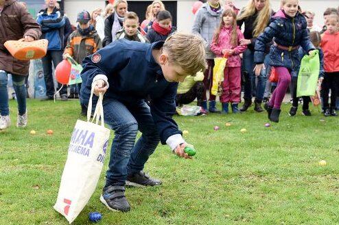Ein Junge hebt ein buntes Ei vom Boden auf, während Kinder und Erwachsene im Hintergrund an einer Eier-Suche teilnehmen.