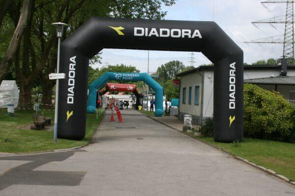 Zwei gro&szlig;e, aufblasbare Startb&ouml;gen mit den Logos &bdquo;DIADORA&ldquo; und &bdquo;RUNNERS POINT&ldquo; stehen auf einem Weg in einem Park.