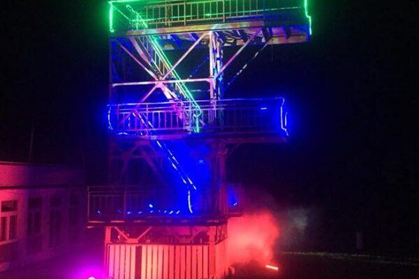 Beleuchtete Stahlkonstruktion mit Treppen und Plattformen, umgeben von Nebel, bei Nacht.