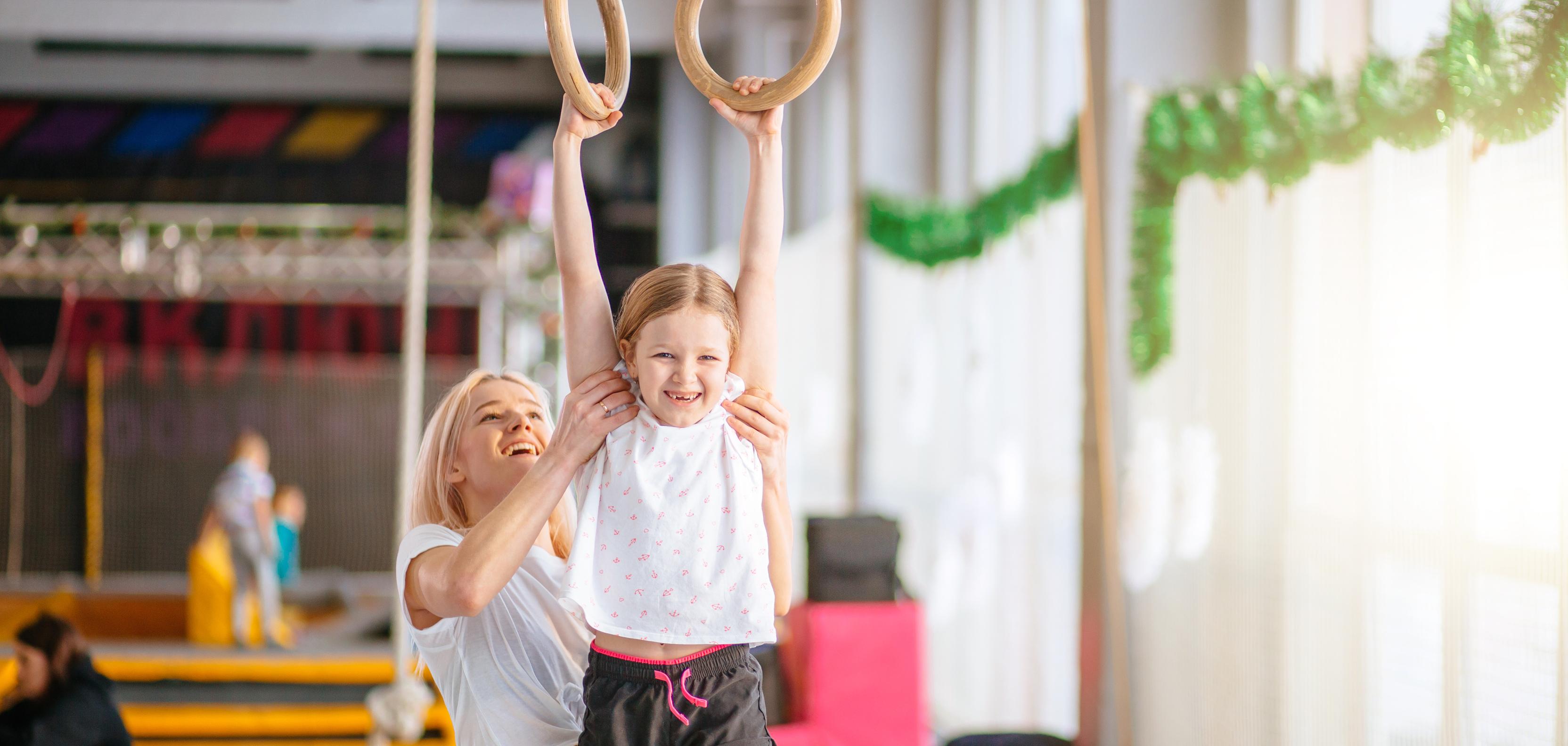 Ein Mädchen hängt an Turnringen, während eine Trainerin sie von unten unterstützt. Heller Turnraum im Hintergrund.