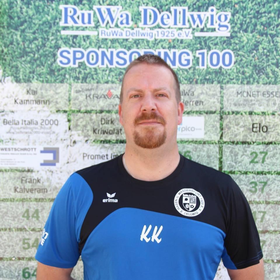 Mann mit kurzem Bart, im schwarzen und blauen Sporttrikot, steht vor einem Sponsoring-Board mit Vereinsnamen.