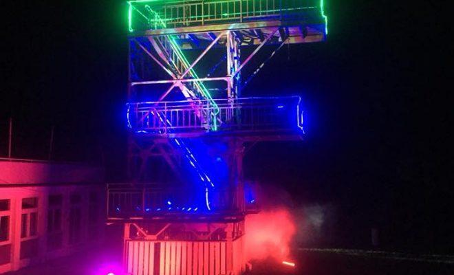 Beleuchtete Stahlkonstruktion mit Treppen und Plattformen, umgeben von Nebel, bei Nacht.