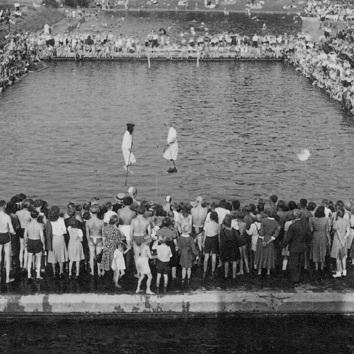 Zuschauer stehen am Rand eines Schwimmbeckens, w&auml;hrend zwei Akteure im Wasser agieren. Historisches Schwarz-Wei&szlig;-Bild.