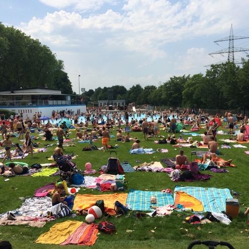 Menschen entspannen auf Liegen im Gras eines &uuml;berf&uuml;llten Schwimmbads an einem sonnigen Tag.