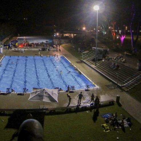Beleuchtetes Schwimmbad mit Menschen bei Nacht, umgeben von B&auml;umen und Sitzgelegenheiten auf der Wiese.