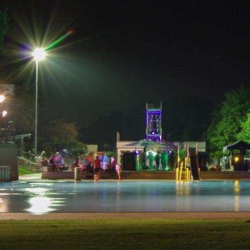 Bunt beleuchteter Schwimmplatz bei Nacht mit Menschen, die sich an der Wasseroberfl&auml;che und im Umfeld aufhalten.