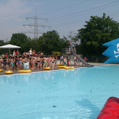 Nahaufnahme eines Schwimmbereichs mit einer Rutsche, umgeben von Kindern und aufblasbaren Booten.