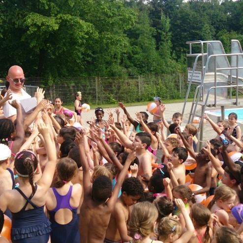 Gruppenspiel von Kindern am Schwimmbad, umgeben von Erwachsenen, mit einem Schwimmlehrer im Vordergrund.