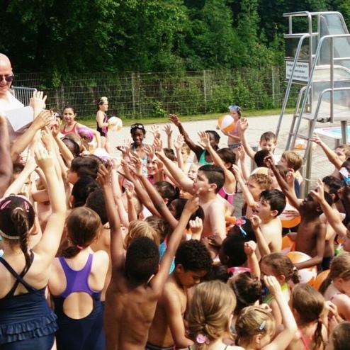 Gruppe begeisterter Kinder umringt einen Erwachsenen am Rand eines Schwimmbeckens in einem Freibad.