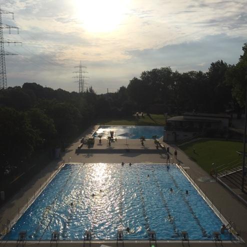 Blick auf ein Freibad mit mehreren Schwimmbecken und Badeg&auml;sten, umgeben von B&auml;umen und einem bew&ouml;lkten Himmel.