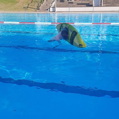 Person in einem gelben Kajak, kopf&uuml;ber im blauen Wasser eines Schwimmbads, mit Wasseroberfl&auml;che und Umgebung.