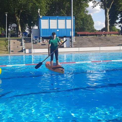 Person paddelt auf einem Stand-Up-Paddle-Board in einem Schwimmbad mit blauem Wasser und B&auml;umen im Hintergrund.