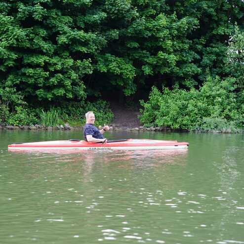Eine Person paddelt in einem roten Kajak auf einem gr&uuml;nen Wasserweg, umgeben von B&auml;umen und Ufervegetation.