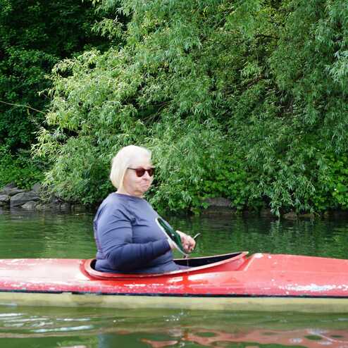 Frau mit blonden Haaren in Sonnenbrille paddelt in rotem Kajak auf einem Gew&auml;sser, umgeben von gr&uuml;ner Vegetation.