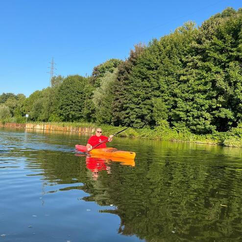 Mann in rotem Shirt paddelt in orangefarbenem Kajak auf ruhigem Wasser, umgeben von gr&uuml;ner Ufervegetation.