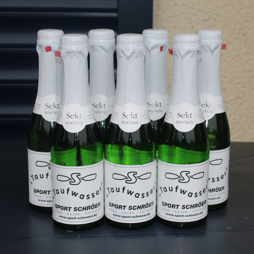 Acht Flaschen Sekt mit den Etiketten &bdquo;Sekt&ldquo; und &bdquo;Taufwasser&ldquo; auf einem dunklen Tisch.