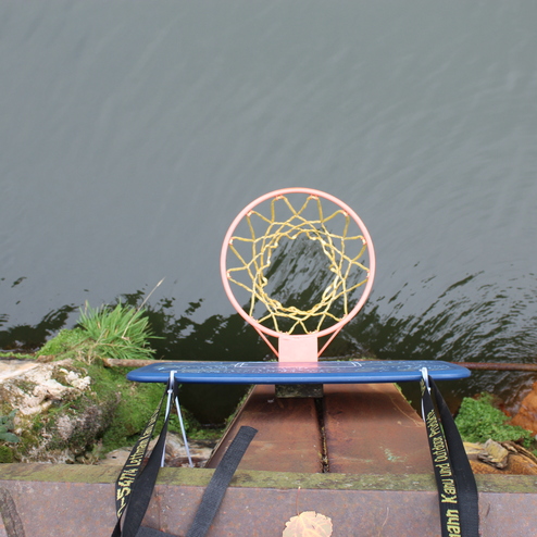 Basketballkorb mit Netz &uuml;ber einem Gew&auml;sser, umgeben von rostigem Metall und gr&uuml;nem Bewuchs.