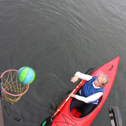Frau im roten Kanu versucht, einen Ball in einen Korb &uuml;ber dem Wasser zu werfen.