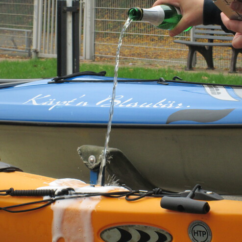 Ein Getr&auml;nk wird aus einer Flasche in ein orangefarbenes Kajak gegossen, im Hintergrund ein blauer Kajak mit Aufschrift.