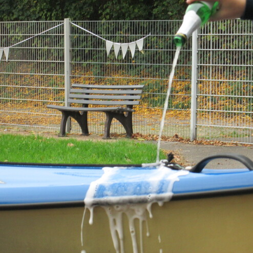 Ein Mann gie&szlig;t Bier auf ein Boot, w&auml;hrend Schaum &uuml;ber den Rand l&auml;uft; im Hintergrund steht eine Bank und Wimpel sind sichtbar.