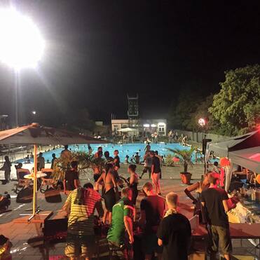 Nachtaufnahme einer Poolparty mit Gästen am Beckenrand, beleuchtet von Scheinwerfern und umgeben von Zelten.