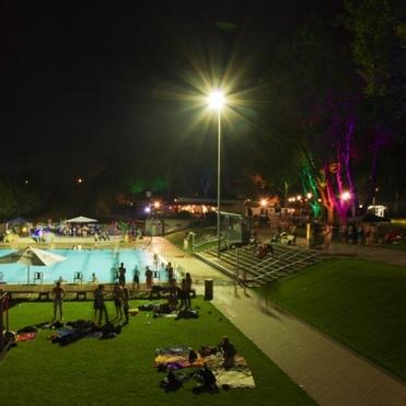 Nachtaufnahme eines beleuchteten Schwimmbeckens mit Menschen am Rand und bunten Lichtern im Hintergrund.