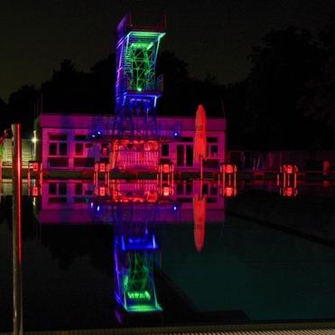 Beleuchteter Sprungturm und Umkleidegeb&auml;ude am Schwimmbecken bei Nacht, reflektiert im ruhigen Wasser.