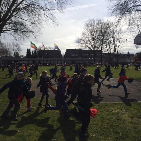 Gruppierung von Kindern in bunter Kleidung, die auf einem Schulhof rennen, um ein Spiel zu spielen. Im Hintergrund sind B&auml;ume und Geb&auml;ude.