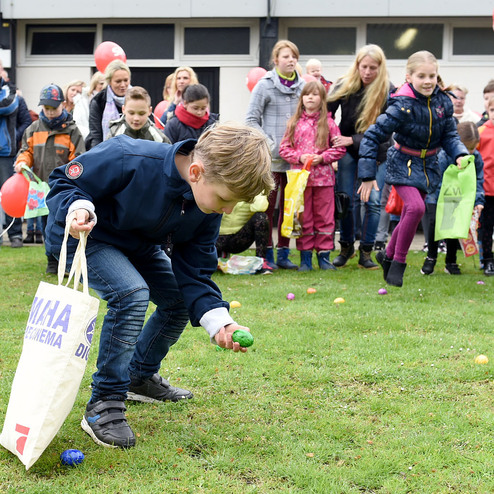 Ein Junge beugt sich nach vorn, um bunte Eier auf einer Rasenfl&auml;che zu sammeln, umgeben von anderen Kindern und Erwachsenen.