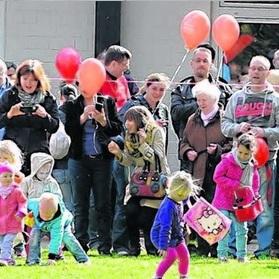 Menschenmenge mit Luftballons beobachtet spielende Kinder auf einer Wiese in sonniger Umgebung.