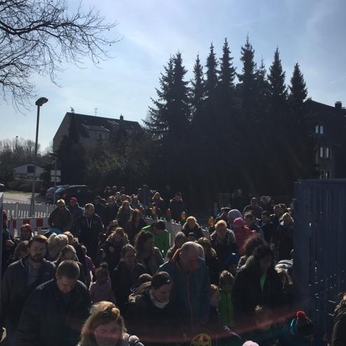 Gro&szlig;e Menschenmenge steht in der Sonne vor einem Zaun, umgeben von B&auml;umen und H&auml;usern im Hintergrund.