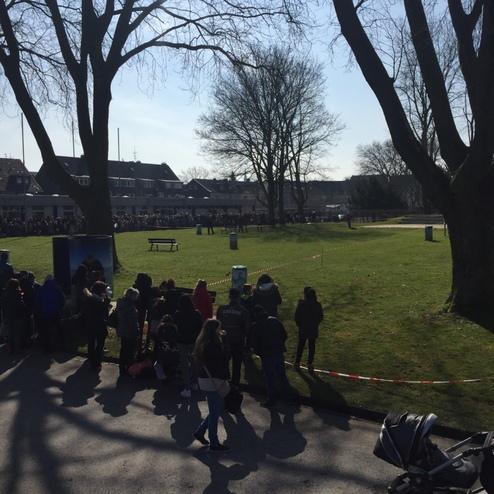 Menschen stehen in einer Schlange auf einer Wiese, umgeben von B&auml;umen, bei sonnigem Wetter.