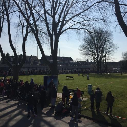 Menschenmenge steht an einem sonnigen Tag in einem Park, umgeben von B&auml;umen, hinter einem benutzten Tisch.