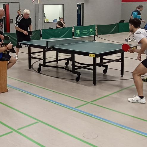 Gruppenspiel von sechs Personen am Tischtennistisch, ein Spieler sitzt auf einer Bank und beobachtet.