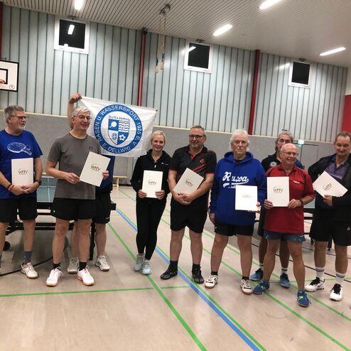 Gruppe von Tischtennisspielern mit Urkunden in einer Halle, im Hintergrund ein Tischtennistisch und ein Banner.