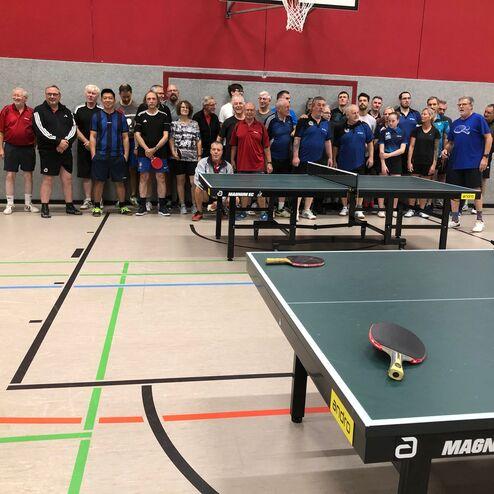 Gruppe von Tischtennisspielern auf einer Sporthalle mit zwei Tischtennisplatten im Vordergrund.