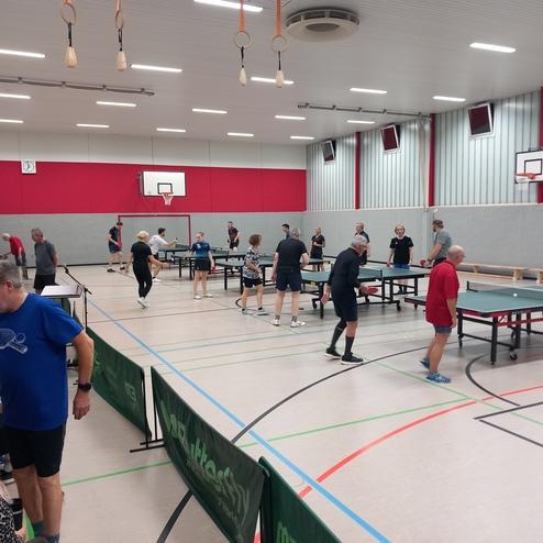 Tischtennisspieler trainieren in einer Sporthalle mit mehreren Tischen und Zuschauern am Rand.