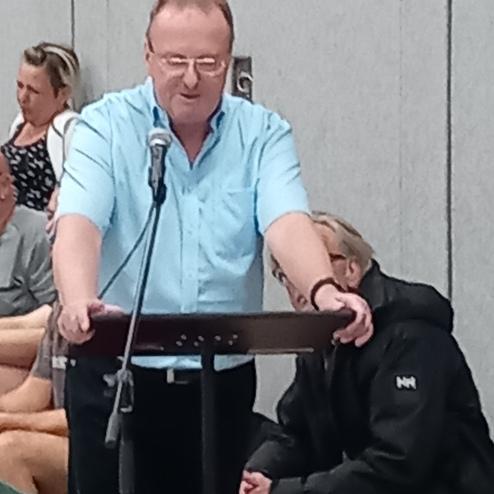 Mann spricht am Mikrofon von einem Tisch, während Zuschauer im Hintergrund sitzen.