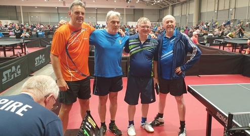 Vier Tischtennisspieler stehen gemeinsam am Tisch, umgeben von weiteren Spielern in einer Halle mit mehreren Tischen.