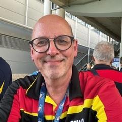 Portrait eines l&auml;chelnden Mannes mit Brille in einem schwarz-rot-gelben Shirt, im Hintergrund verschwommene Personen.