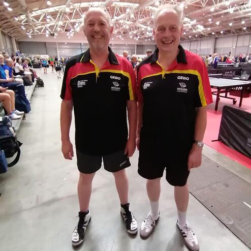 Zwei Tischtennisspieler in einheitlichen Trikots stehen l&auml;chelnd in einer Sporthalle mit mehreren Tischtennisplatten.