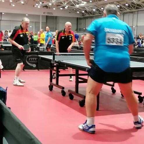 Zwei Tischtennisspieler in Aktion an einem Tisch, im Hintergrund weitere Spieler und Zuschauer in einem Turniersaal.