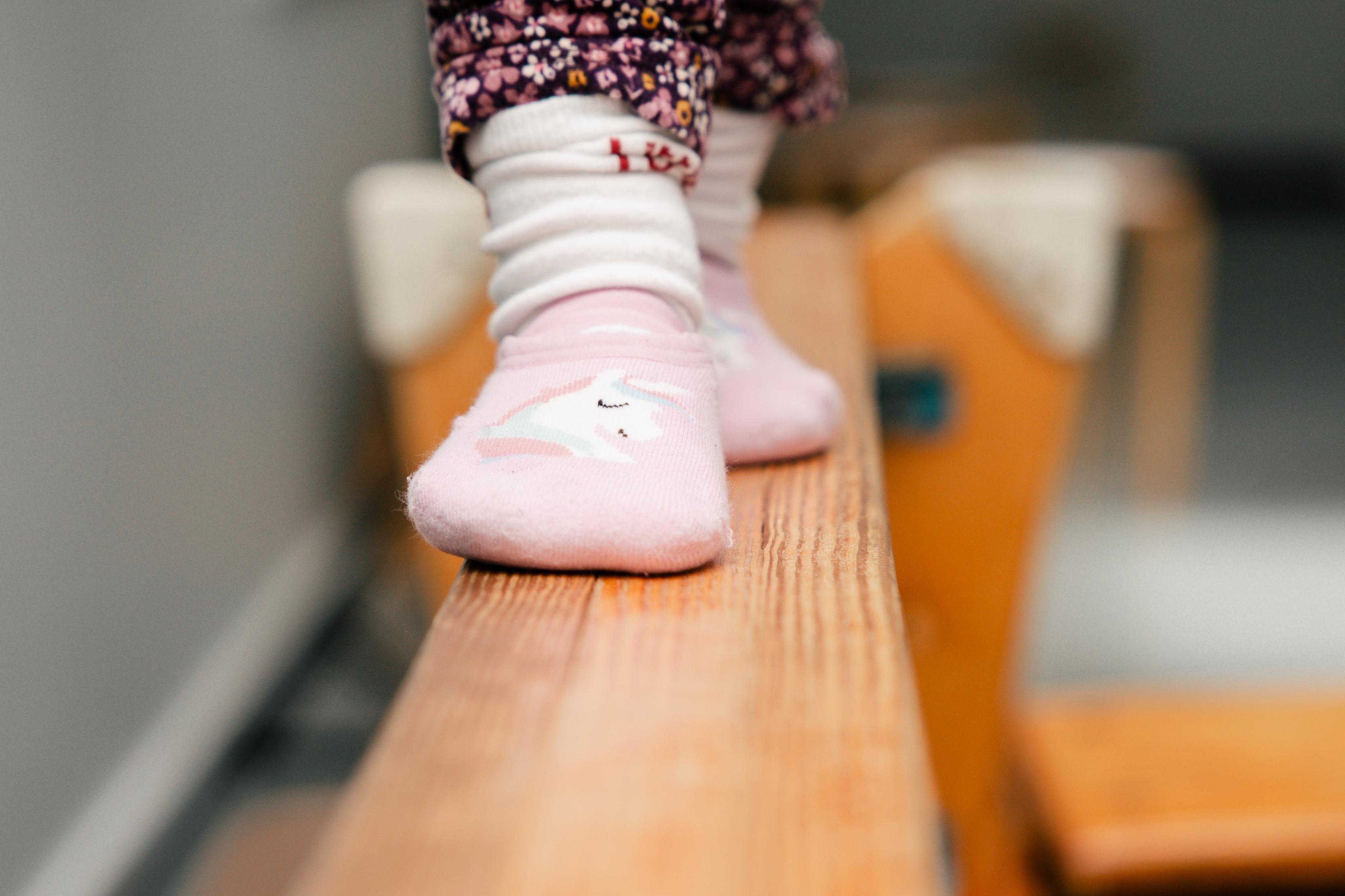 Ein kleines Kind balanciert auf einem niedrigem Holzbrett in rosa Socken mit Blumendruck.