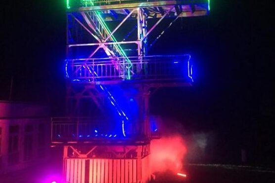 Beleuchtete Stahlkonstruktion mit Treppen und Plattformen, umgeben von Nebel, bei Nacht.