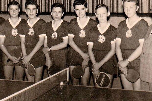 Tischtennis-Jugendmannschaft von RuWa Dellwig mit Betreuern, historische Aufnahme Historisches Schwarz-Weiß-Foto: acht Jugendliche in dunklen Trikots mit Vereinswappen halten Tischtennisschläger. Neben ihnen stehen vier Männer im Anzug, im Vordergrund ist eine Tischtennisplatte mit Netz zu sehen.