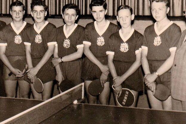 Historisches Schwarz-Wei&szlig;-Foto: acht Jugendliche in dunklen Trikots mit Vereinswappen halten Tischtennisschl&auml;ger. Neben ihnen stehen vier M&auml;nner im Anzug, im Vordergrund ist eine Tischtennisplatte mit Netz zu sehen.