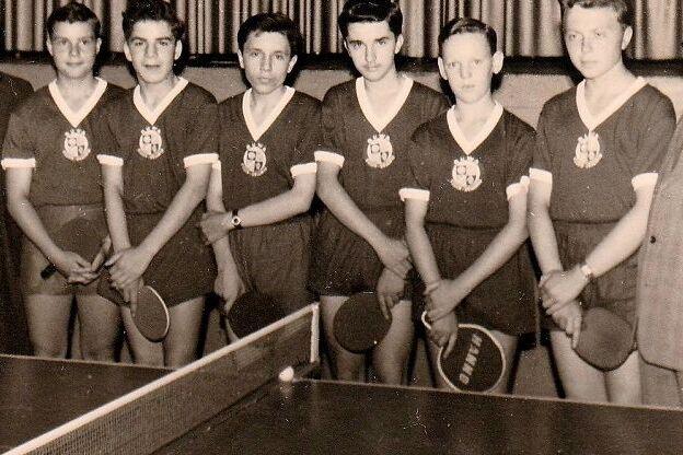 Historisches Schwarz-Wei&szlig;-Foto: acht Jugendliche in dunklen Trikots mit Vereinswappen halten Tischtennisschl&auml;ger. Neben ihnen stehen vier M&auml;nner im Anzug, im Vordergrund ist eine Tischtennisplatte mit Netz zu sehen.