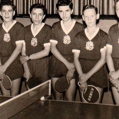Tischtennis-Jugendmannschaft von RuWa Dellwig mit Betreuern, historische Aufnahme Historisches Schwarz-Weiß-Foto: acht Jugendliche in dunklen Trikots mit Vereinswappen halten Tischtennisschläger. Neben ihnen stehen vier Männer im Anzug, im Vordergrund ist eine Tischtennisplatte mit Netz zu sehen.