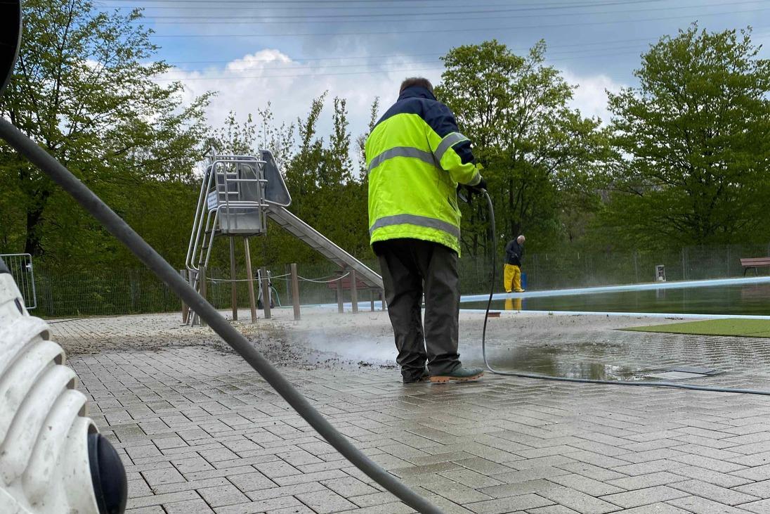 Mann in reflektierender Jacke reinigt mit Hochdruckreiniger eine gepflasterte Fl&auml;che neben einem Spielplatz.